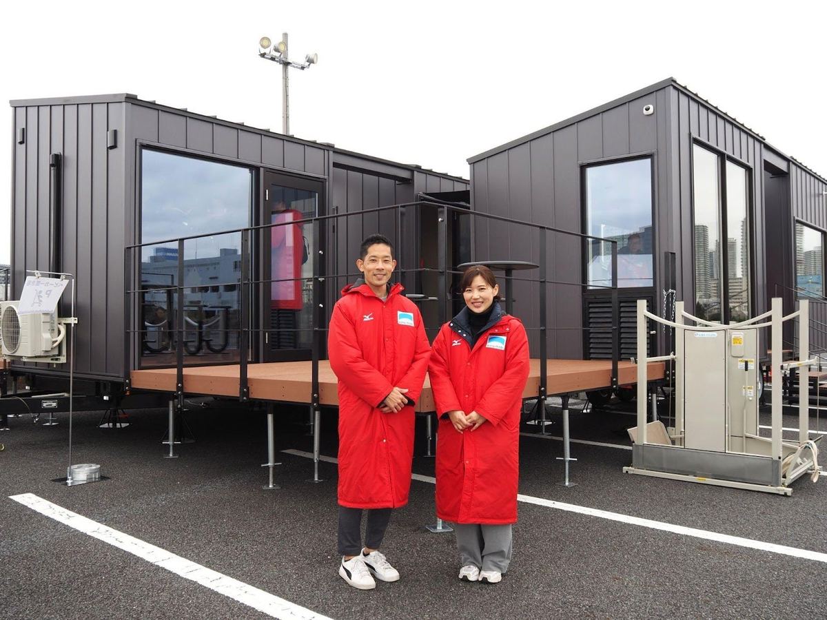 旅客ターミナルとして使われた黒い外壁のトレーラーハウスの前に立つ、赤い上着の2人