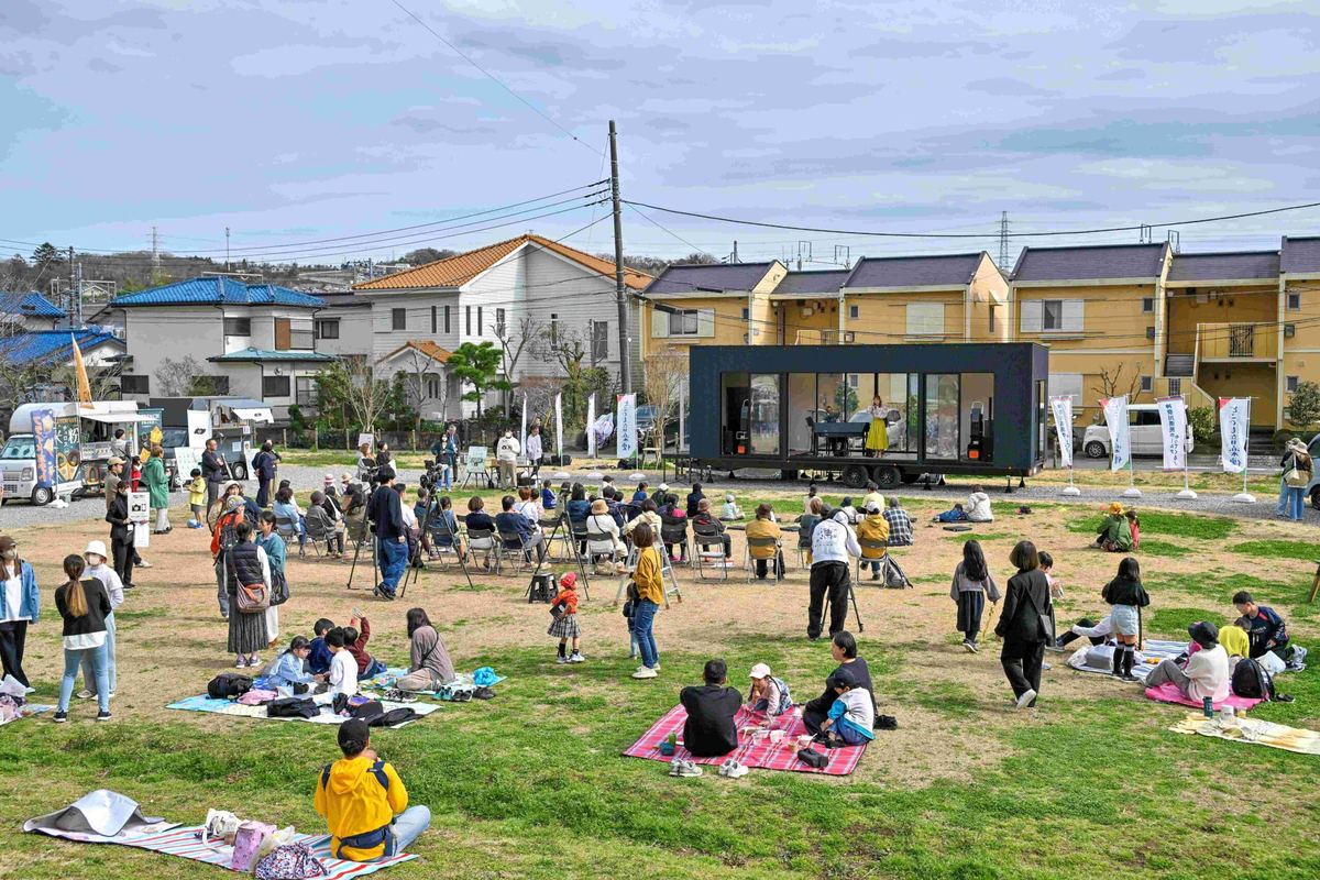 住宅街の芝生広場に置いたトレーラーハウスのステージと、レジャーシートで聴く人たち